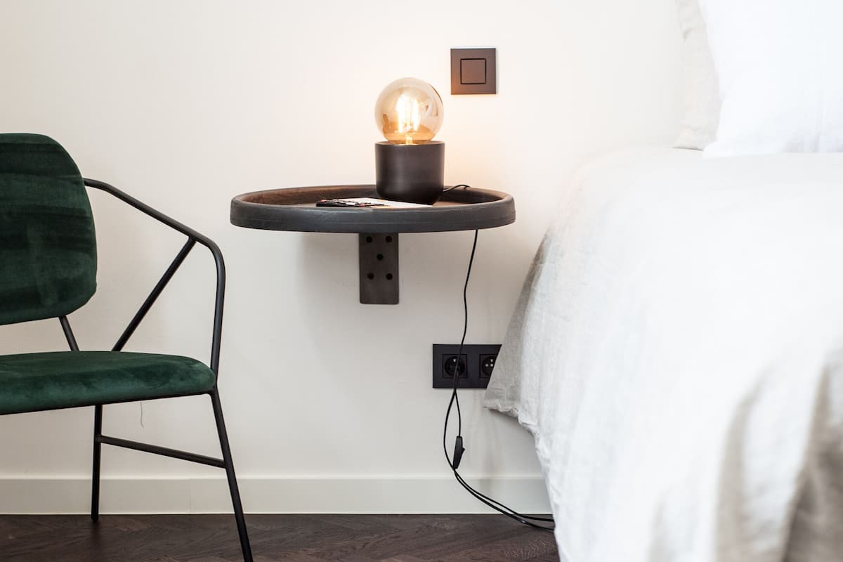 Ein Schlafzimmer mit Bett, Nachttisch mit Lampe, Stuhl und einer Steckdose.