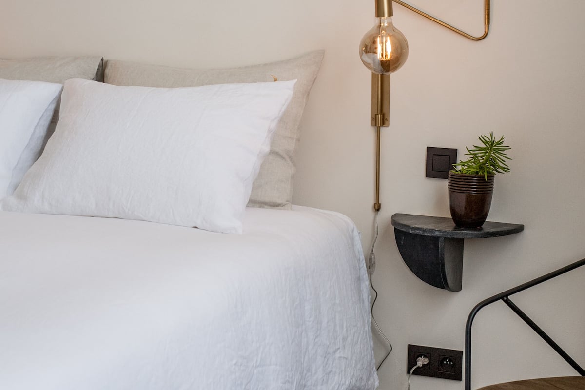 Ein ordentlich bezogenes Bett mit weißer Bettwäsche, eine Wandlampe, ein kleiner Topfpflanze und ein Stuhl.