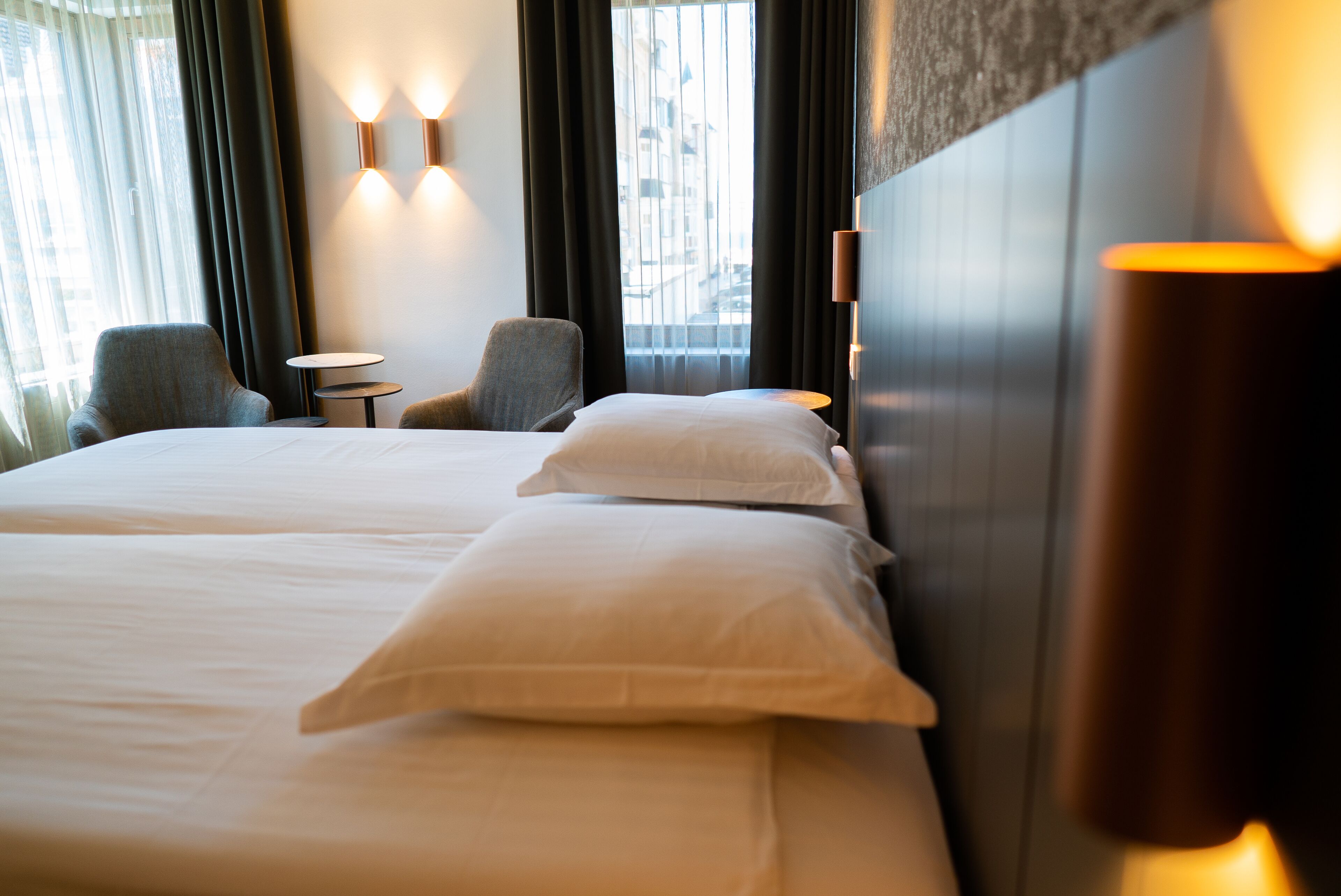 Quarto de hotel com duas camas, uma mesa pequena, duas poltronas e vista para a cidade através de grandes janelas.