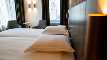 Quarto de hotel com duas camas, uma mesa pequena, duas poltronas e vista para a cidade através de grandes janelas.