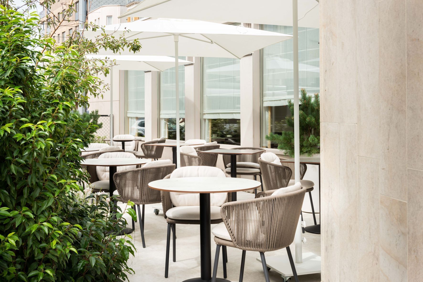 Un área de café al aire libre con mesas redondas y sillas de mimbre, rodeada de paredes de vidrio y follaje.