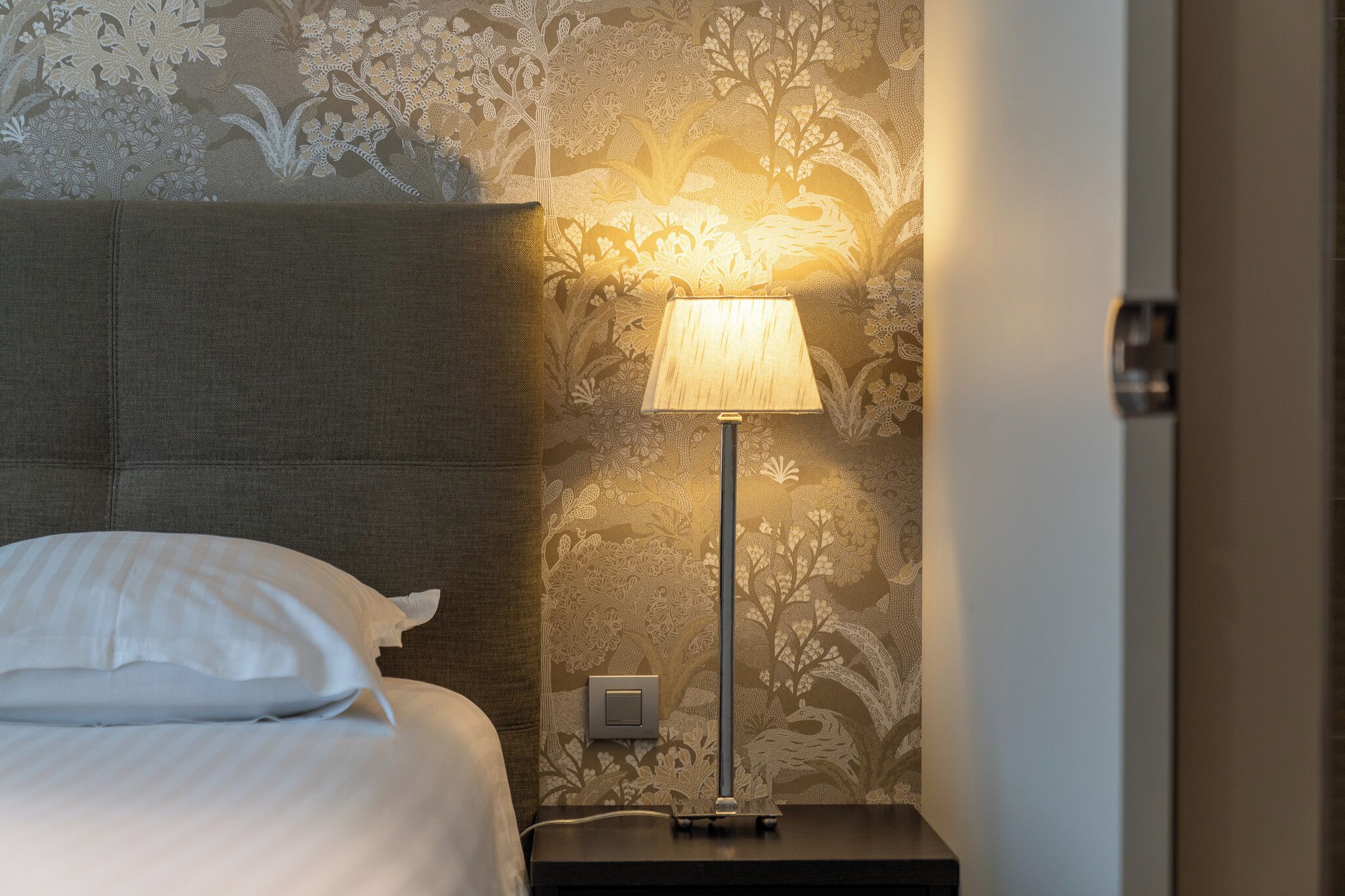 Una cama bien tendida con sábanas blancas, una mesita de noche con lámpara y una pared con estampado floral.