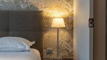 Una cama bien tendida con sábanas blancas, una mesita de noche con lámpara y una pared con estampado floral.