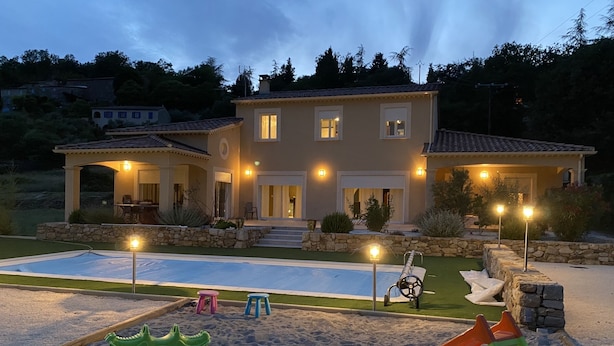 Una casa de dos pisos con piscina, mobiliario exterior y un patio bien iluminado.