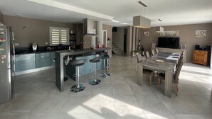 Una cocina moderna con una isla central, taburetes de bar, una zona de comedor con una mesa grande y sillas, un televisor y una escalera que sube a otro nivel.