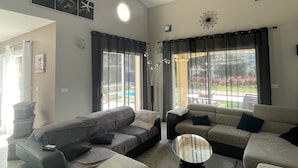 Una sala de estar con un sofá modular grande, una mesa de centro y una chimenea. Hay ventanas amplias con cortinas, un techo con vigas a la vista y un reloj en la pared.