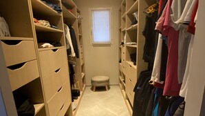 Un armario estrecho con espacio para caminar, múltiples estantes y cajones, un taburete pequeño y una ventana con persianas.