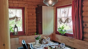 Interior de una cabaña de madera con una mesa de comedor preparada para cuatro personas, sillas, un radiador y ventanas con cortinas.