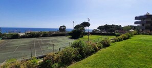 A tennis court with a net, surrounded by greenery and a building in the background. - Cascais by the Sea (Cascais)