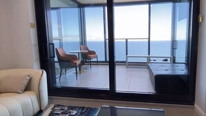 Una habitación moderna con un gran ventanal corredero que da a un balcón con vistas al mar. Hay un sofá y una pequeña mesa con sillas dentro de la habitación.