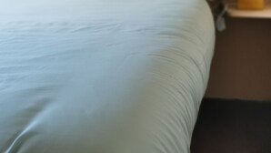 Una cama con un edredón de color claro y almohadas, una mesita de noche con una lámpara y una pared oscura.
