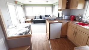 Una cocina compacta con armarios de madera, un fregadero y un lavavajillas. Hay una zona de comedor con una mesa y sillas, y un sofá al fondo.