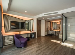 A modern hotel room with a large mirror, a vanity with a sink, a purple armchair, and a desk with a chair.