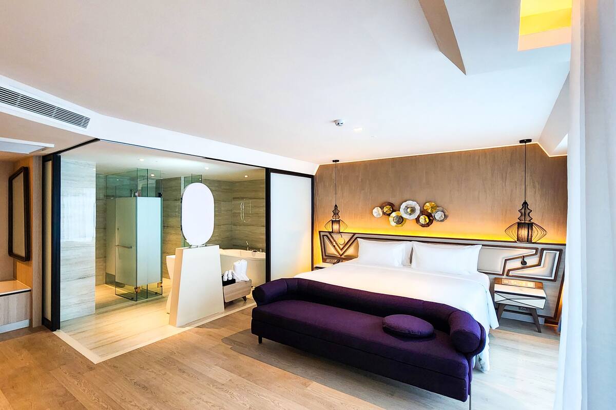 Moderni hotellihuone, jossa on suuri sänky, violetti sohva, lasiseinällä erotettu suihkutila ja puusta tehty korosteseinä.