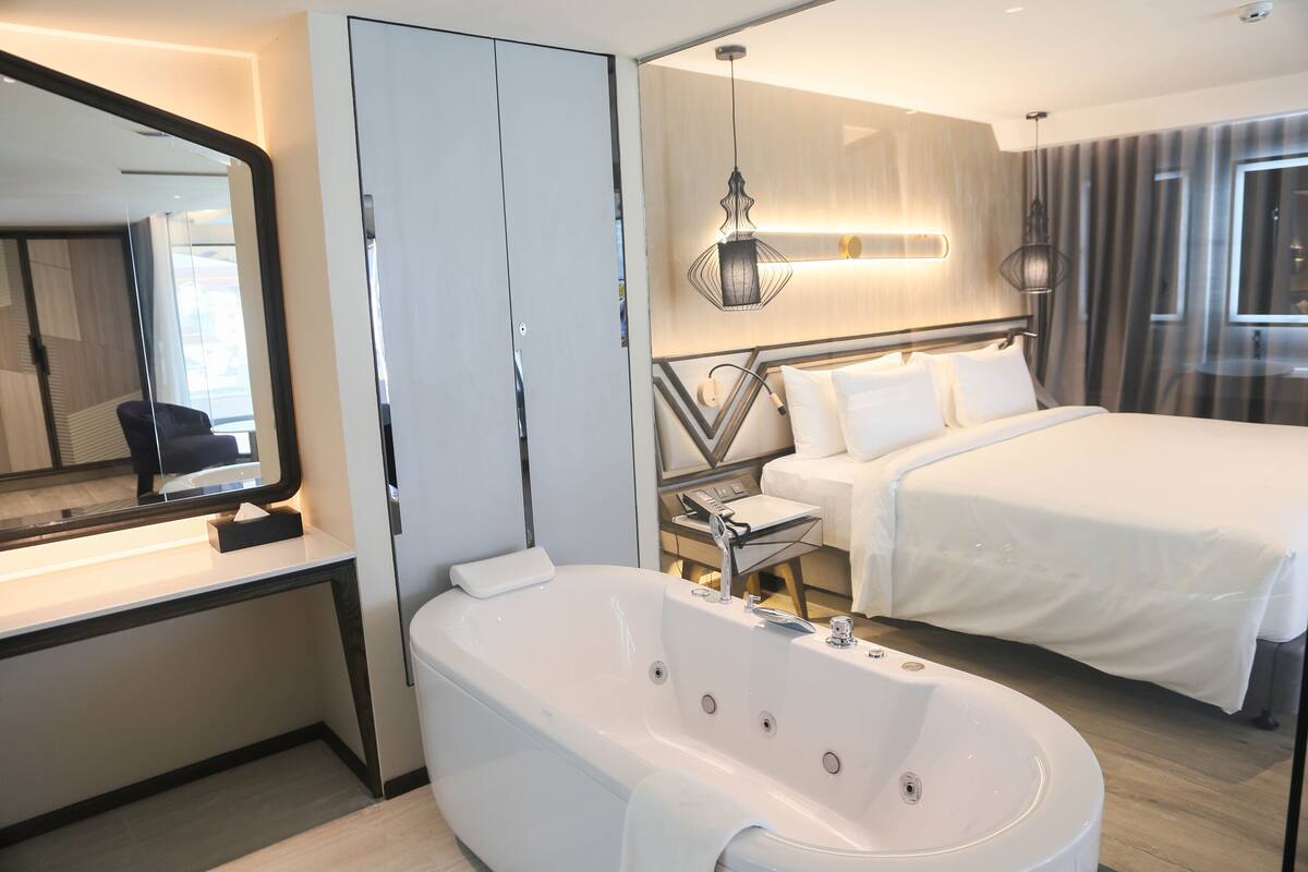 Moderni hotellihuone, jossa on suuri sänky, erillinen kylpyhuone, jossa on kylpyamme, peilillä varustettu meikkipöytä ja yöpöytä.