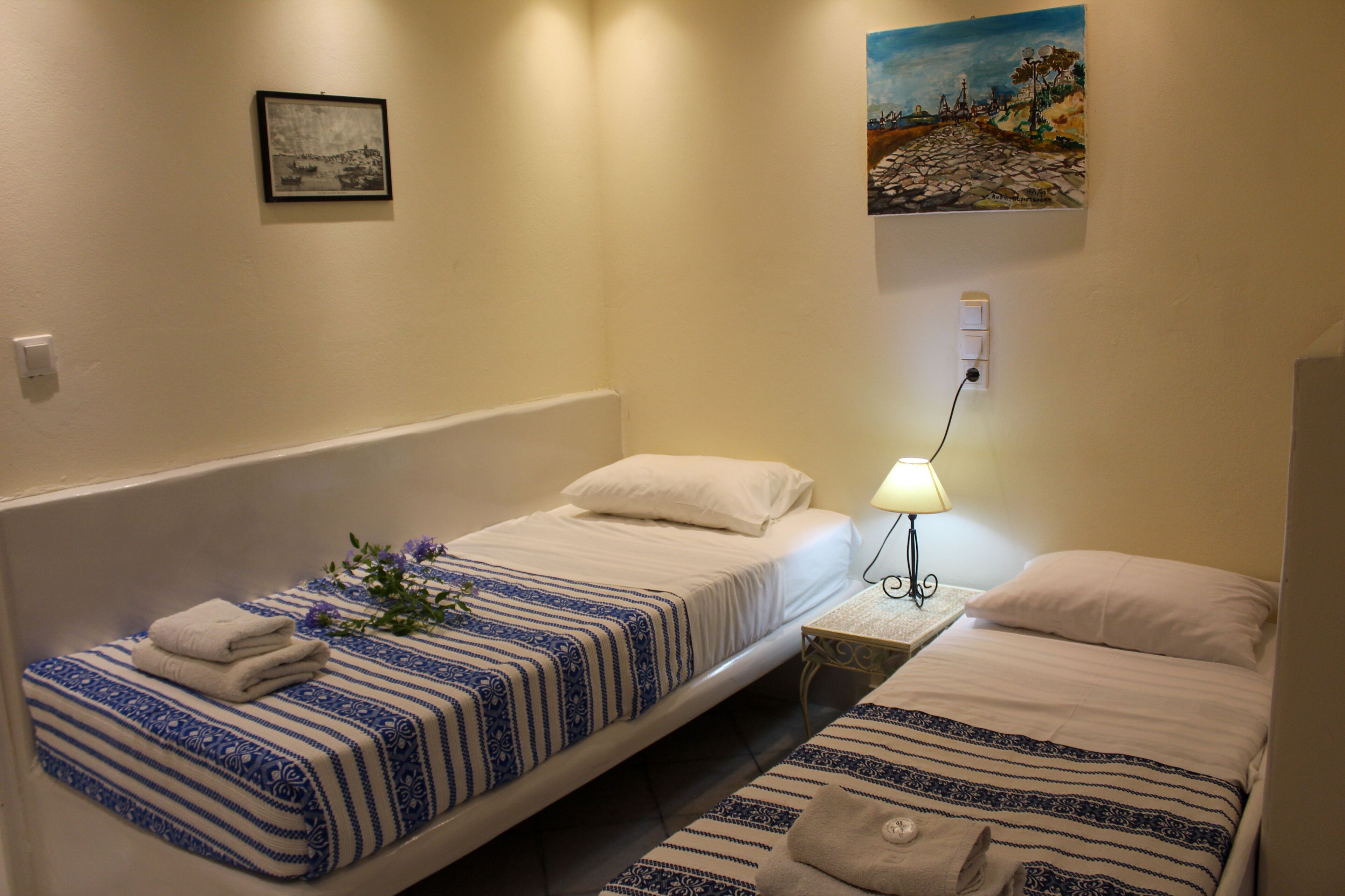 A hotel room with two single beds, each with a blue and white striped bedspread and white pillows. There is a framed picture on the wall above the left bed and a painting above the right bed. A small table with a lamp is between the beds.