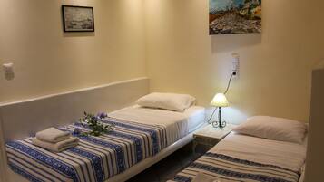 A hotel room with two single beds, each with a blue and white striped bedspread and white pillows. There is a framed picture on the wall above the left bed and a painting above the right bed. A small table with a lamp is between the beds.