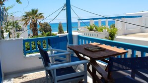 A balcony with a wooden table and blue and white chairs, overlooking a coastal view with palm trees and a clear sky.