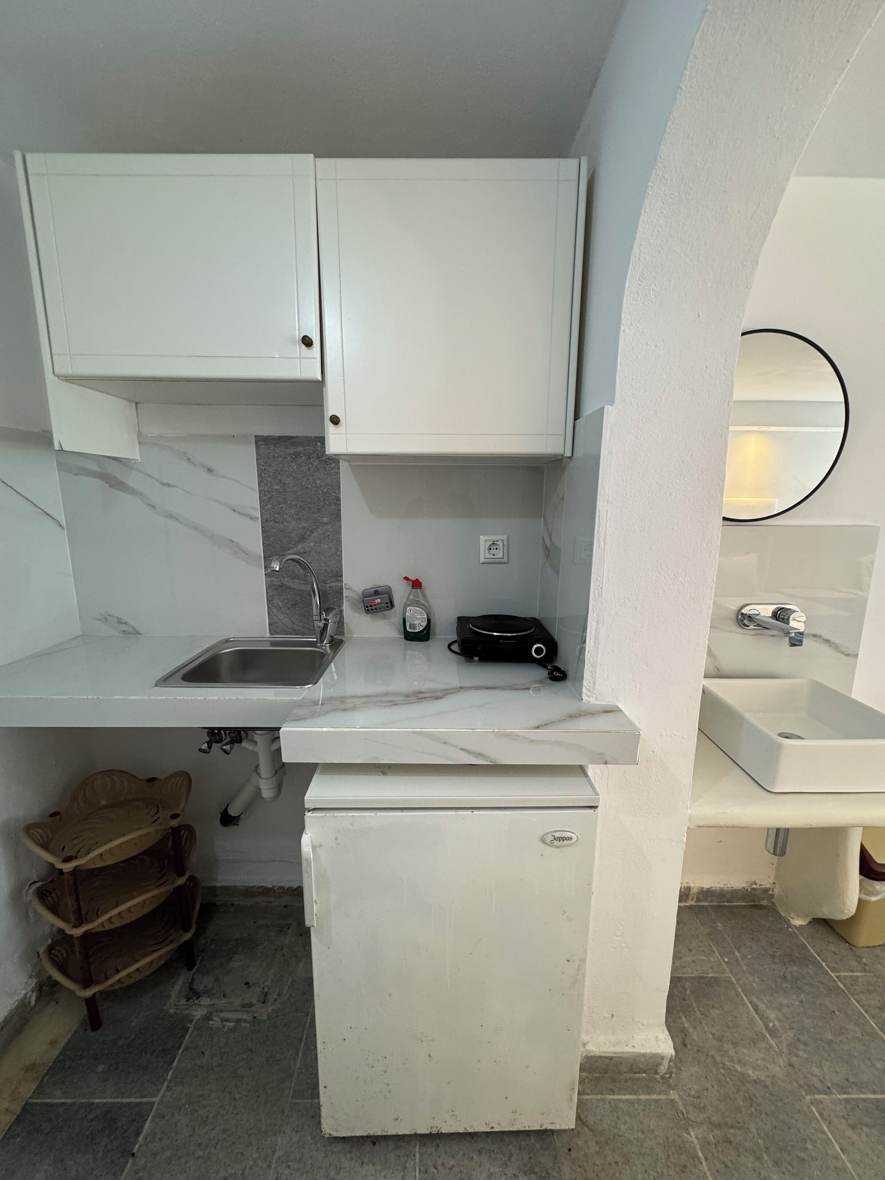 Una cocina compacta con armarios blancos, un lavamanos empotrado, un refrigerador pequeño y una encimera con una tetera y una cafetera.