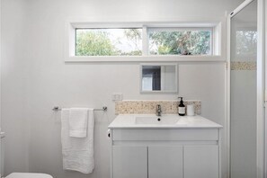 Un baño con un lavamanos blanco, un perchero, una ventana sobre el lavamanos y un espejo.