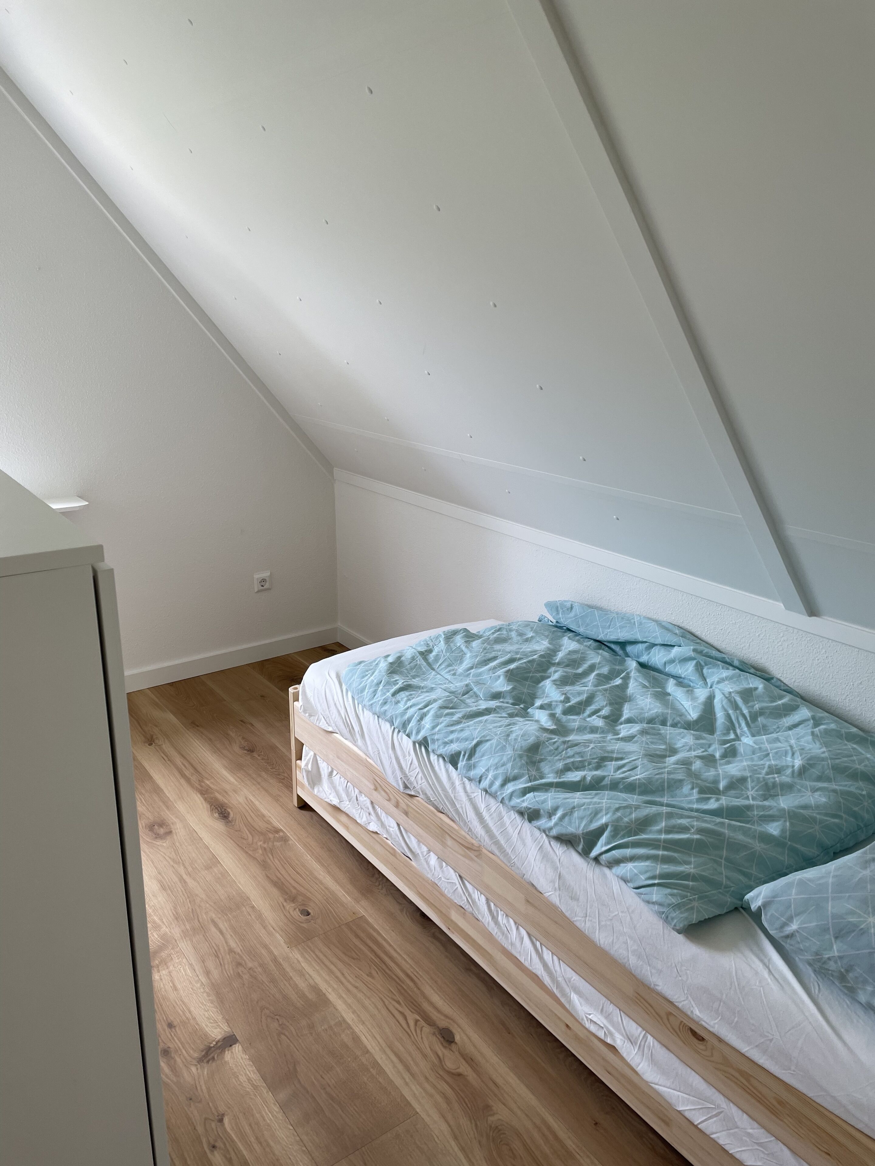 A small, sloped ceiling room with a single bed, wooden flooring, and white walls.