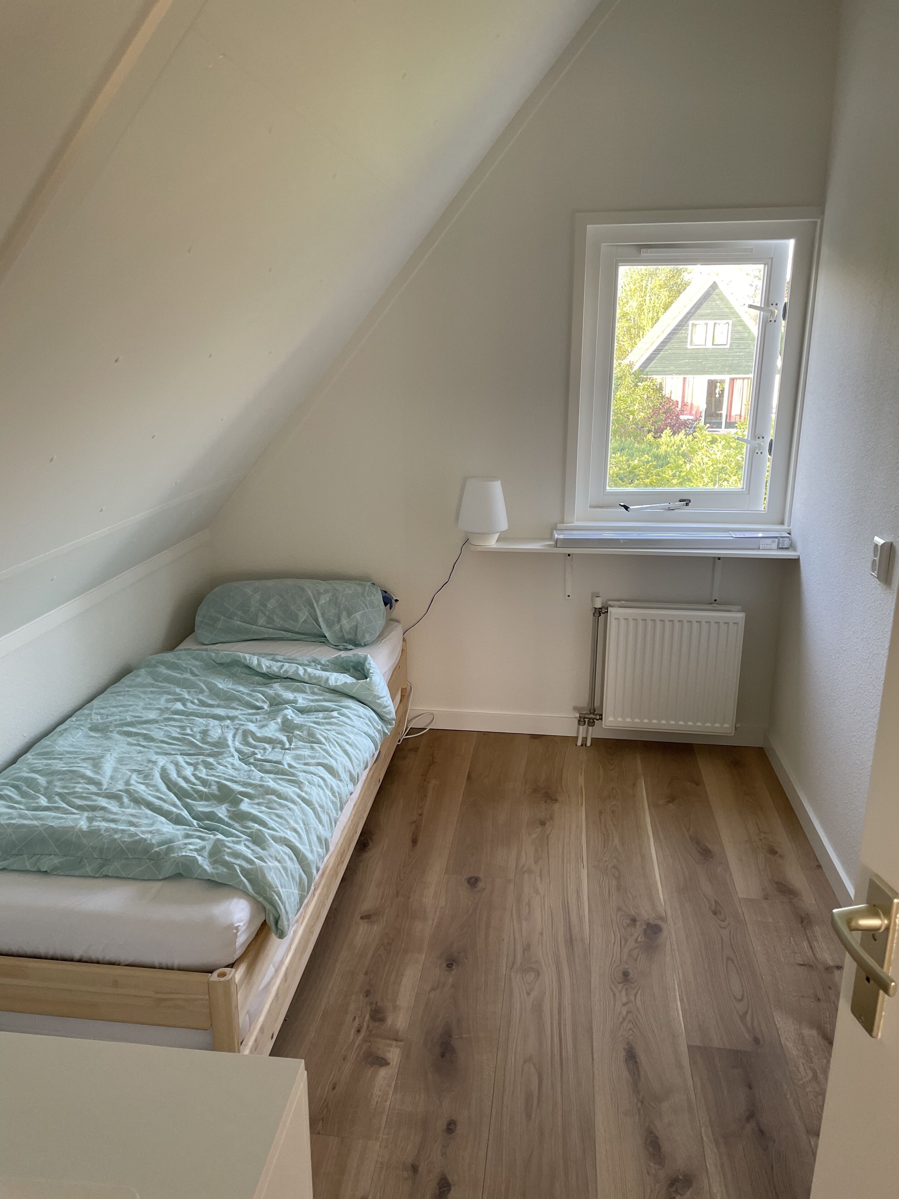 A small, single-bed room with a sloped ceiling, a window with a view of greenery, and a radiator.