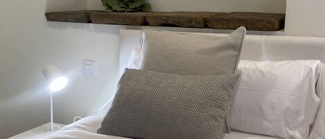 A neatly made bed with white linens, two patterned pillows, a small green plant on a wooden shelf, and a bedside lamp.