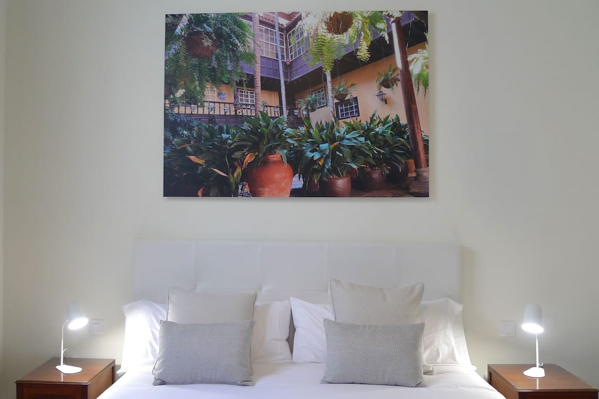 Een hotelkamer met een bed, twee nachtkastjes, een hoofdeinde en een groot schilderij van een binnentuin met planten en een gebouw.