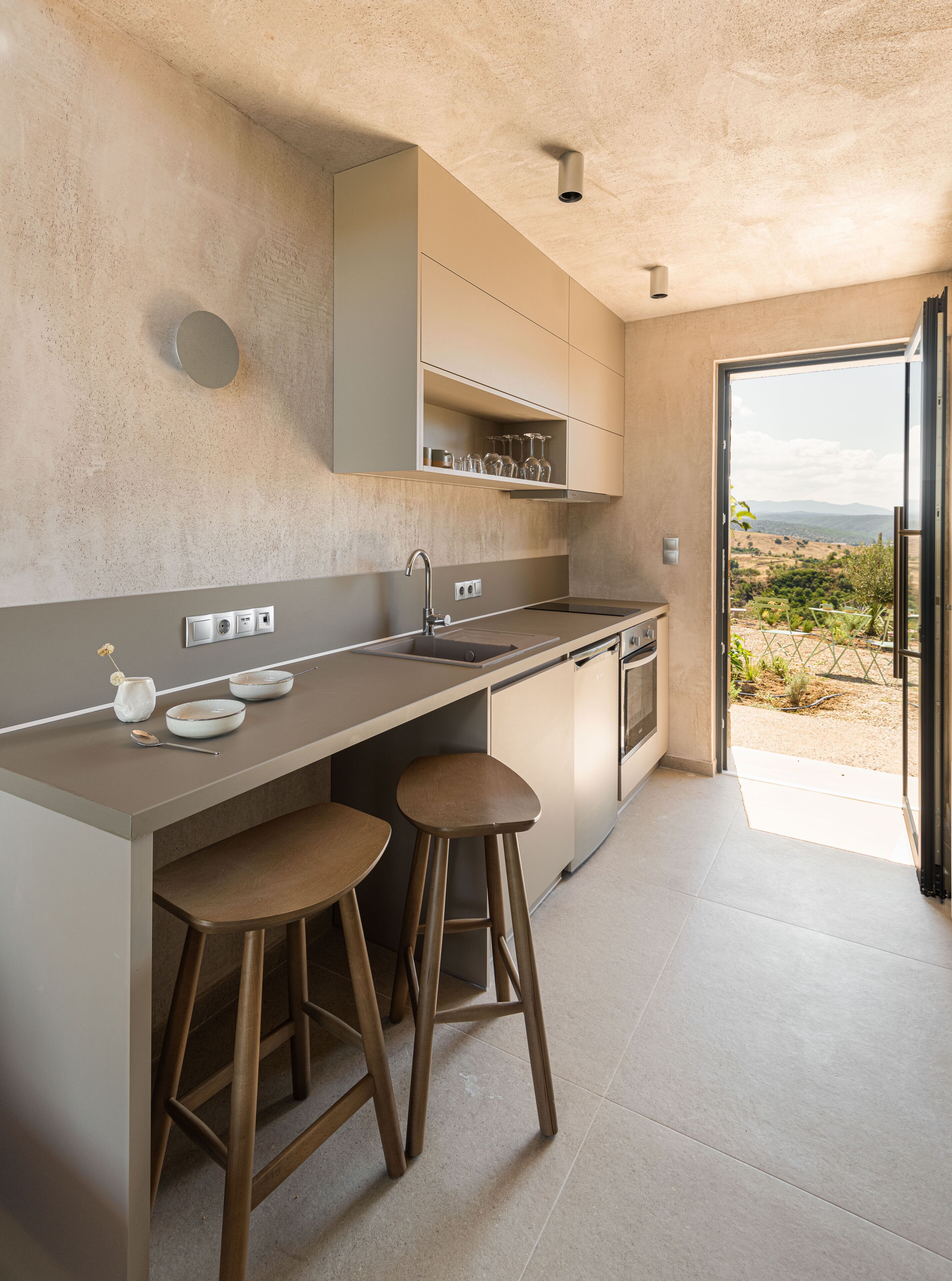 Una cocina moderna con una isla grande, taburetes de bar y vistas al campo a través de una puerta abierta.