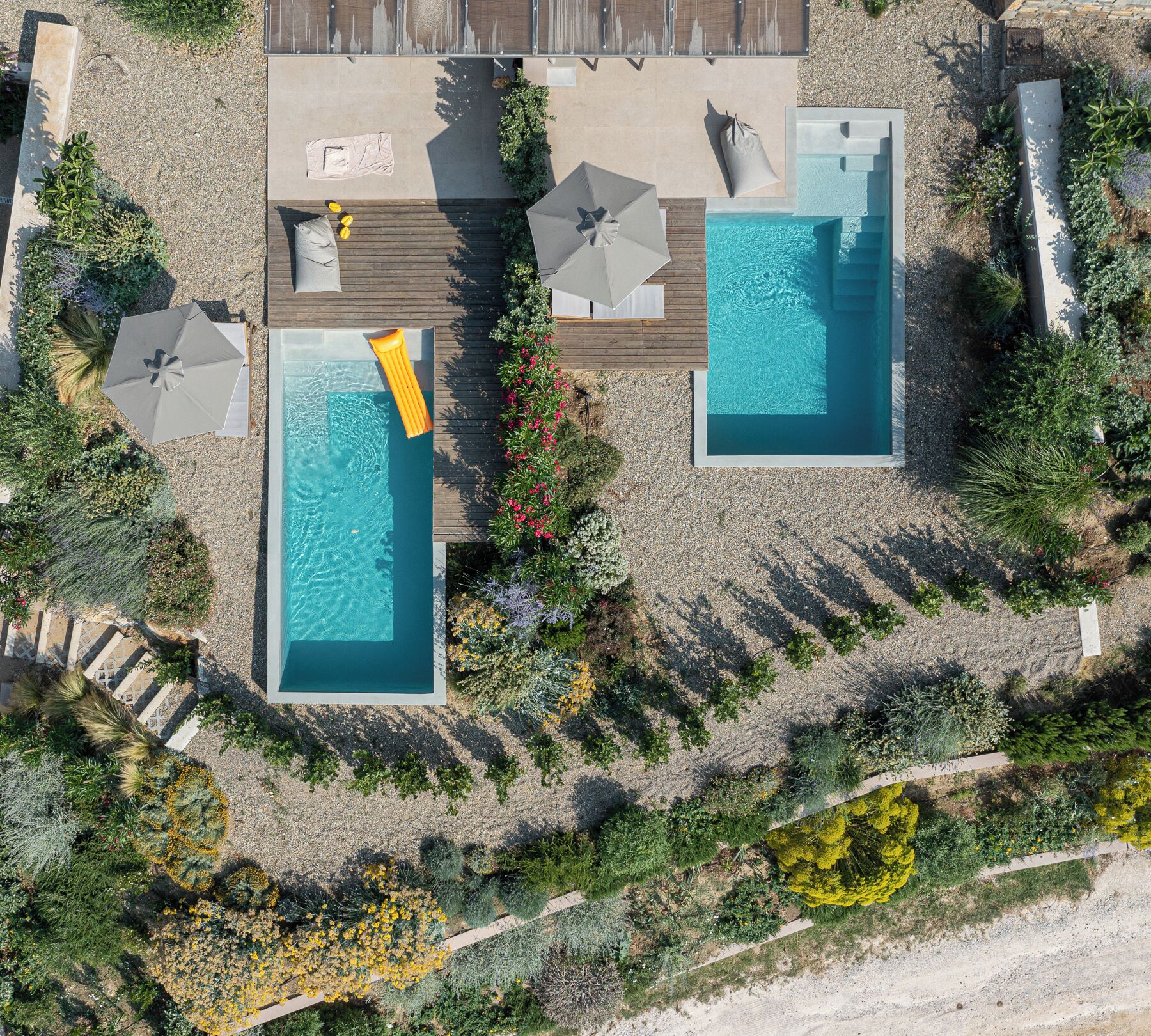 An aerial view of a resort with two rectangular swimming pools, surrounded by wooden decking, and shaded by umbrellas.