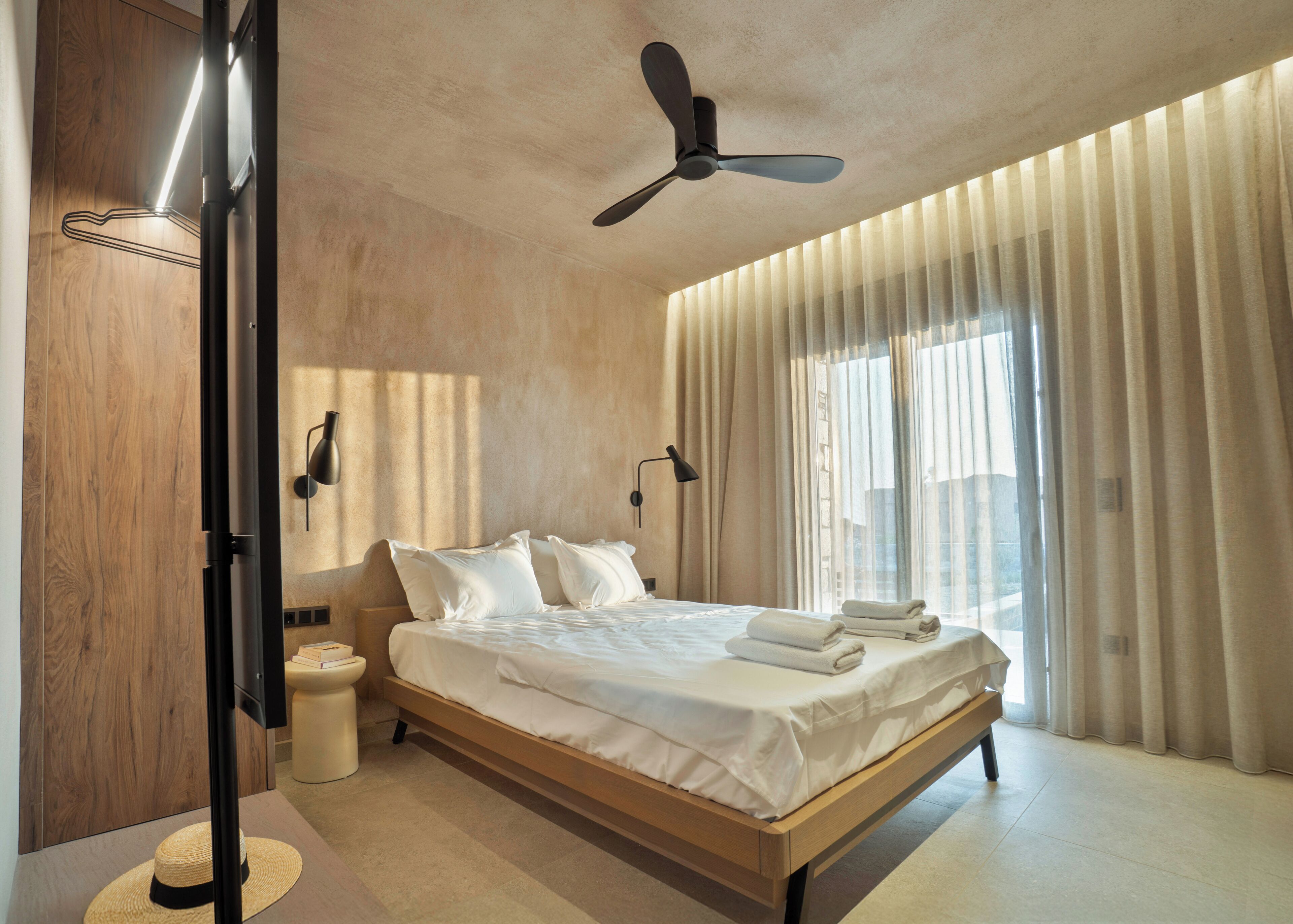 A modern hotel room with a large bed, ceiling fan, and large windows with sheer curtains.