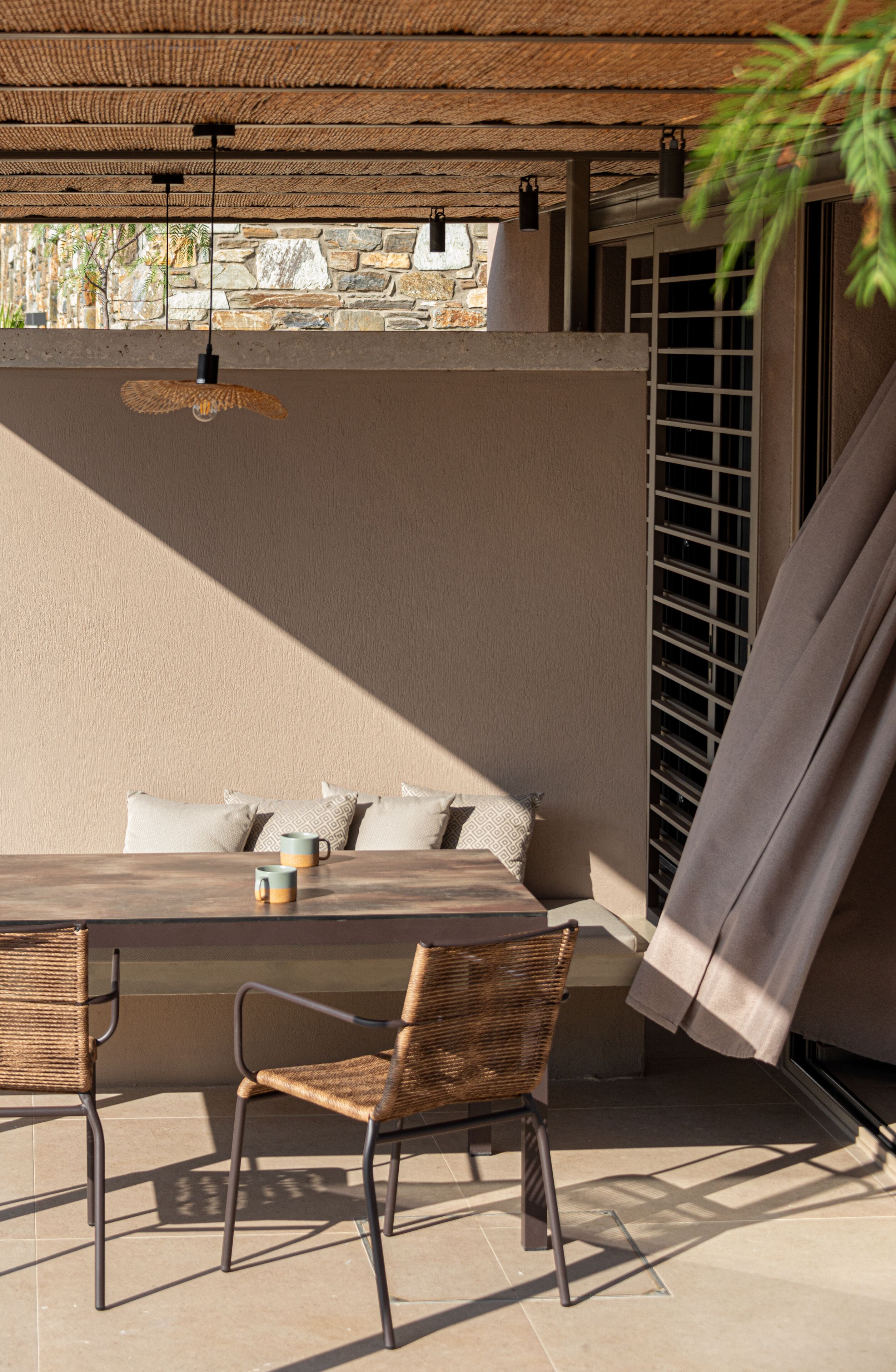 A sunlit outdoor seating area with a stone wall backdrop, a woven chair, and a table with cushions and a mug.