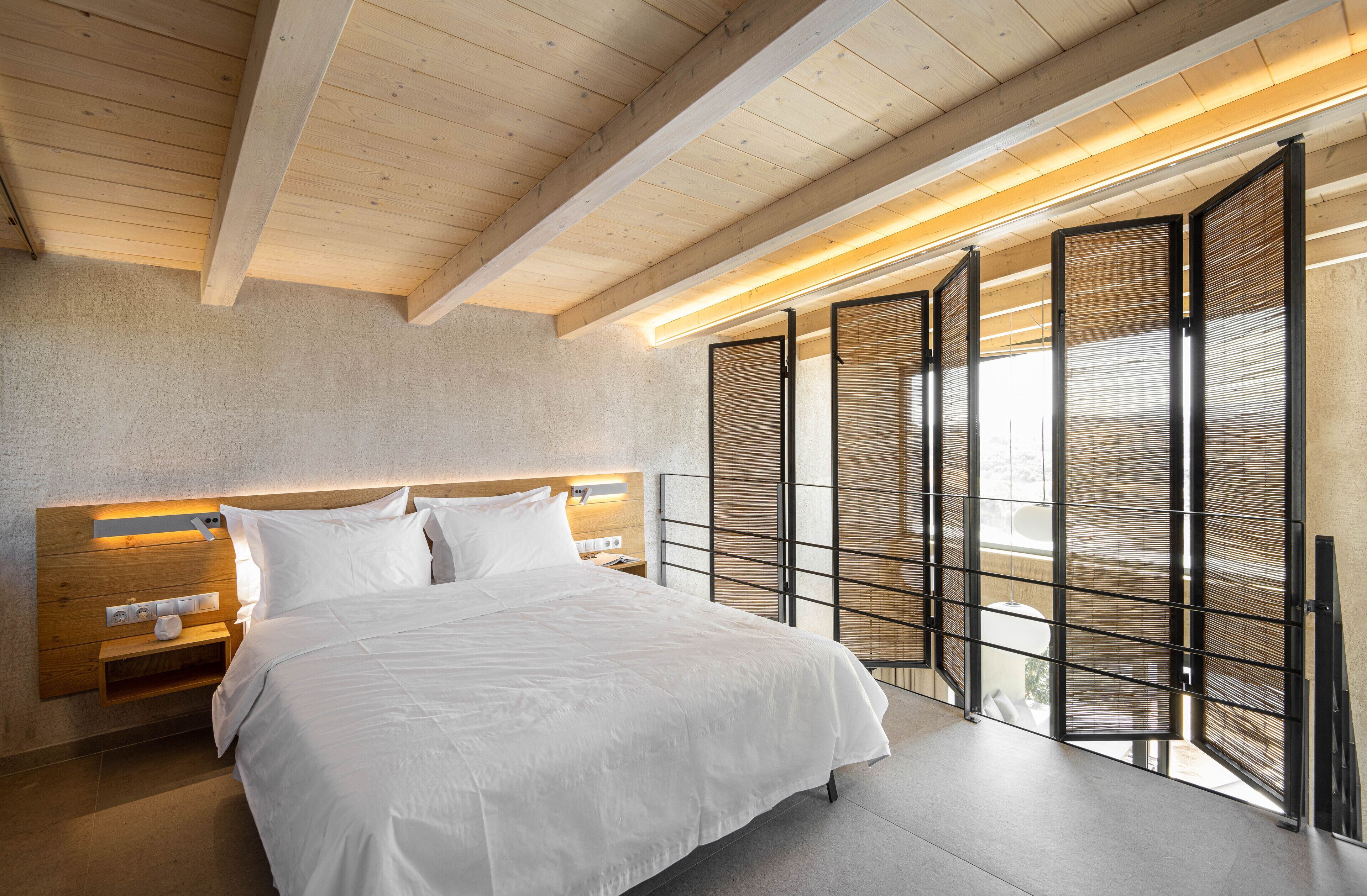 Ett modernt hotellrum med en stor säng, ett träpanelat sänggavel, sängbord och utsikt genom en trävägg.