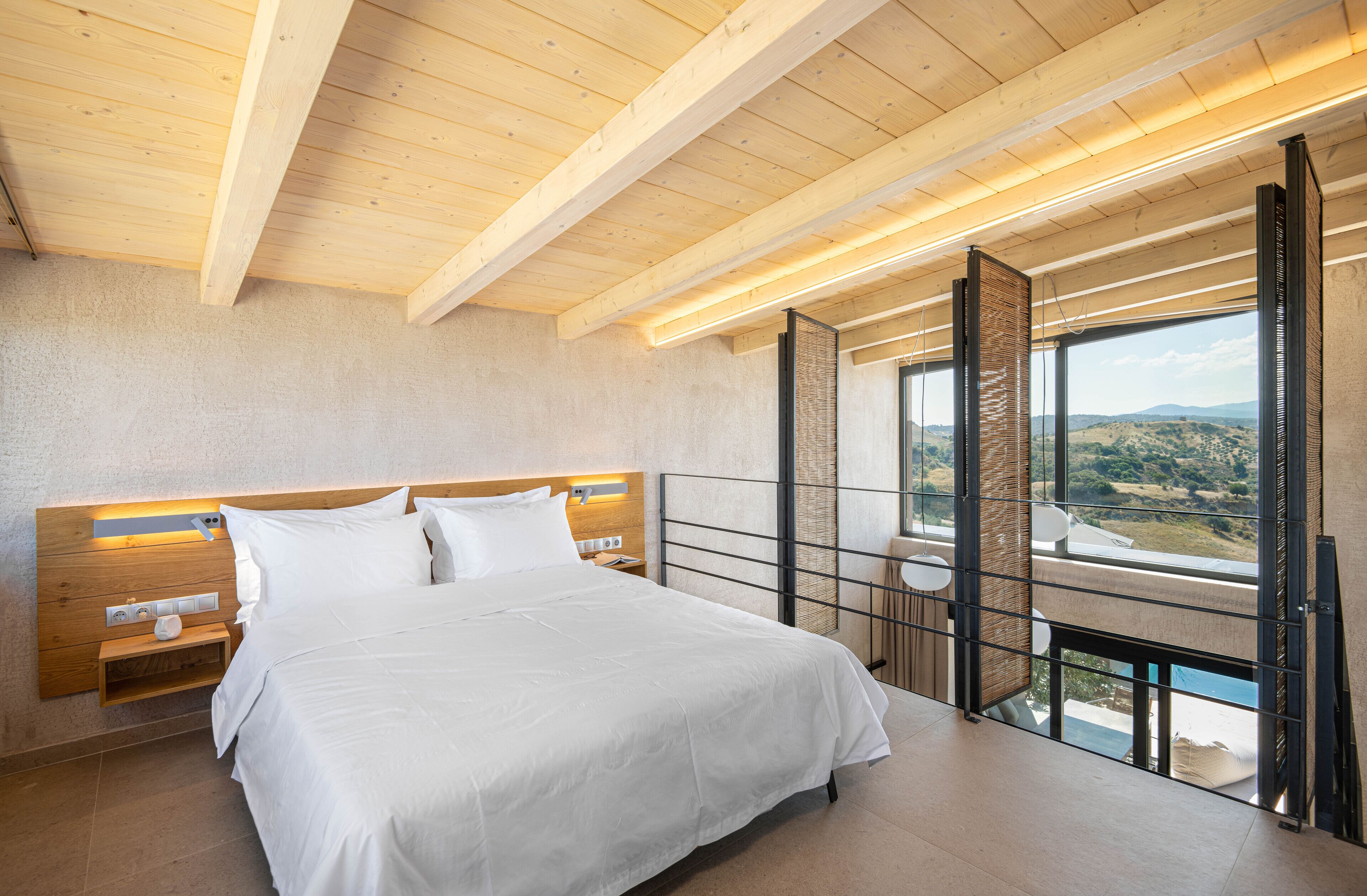 A modern bedroom with a large bed, wooden ceiling beams, a nightstand, and a view of the outdoors.