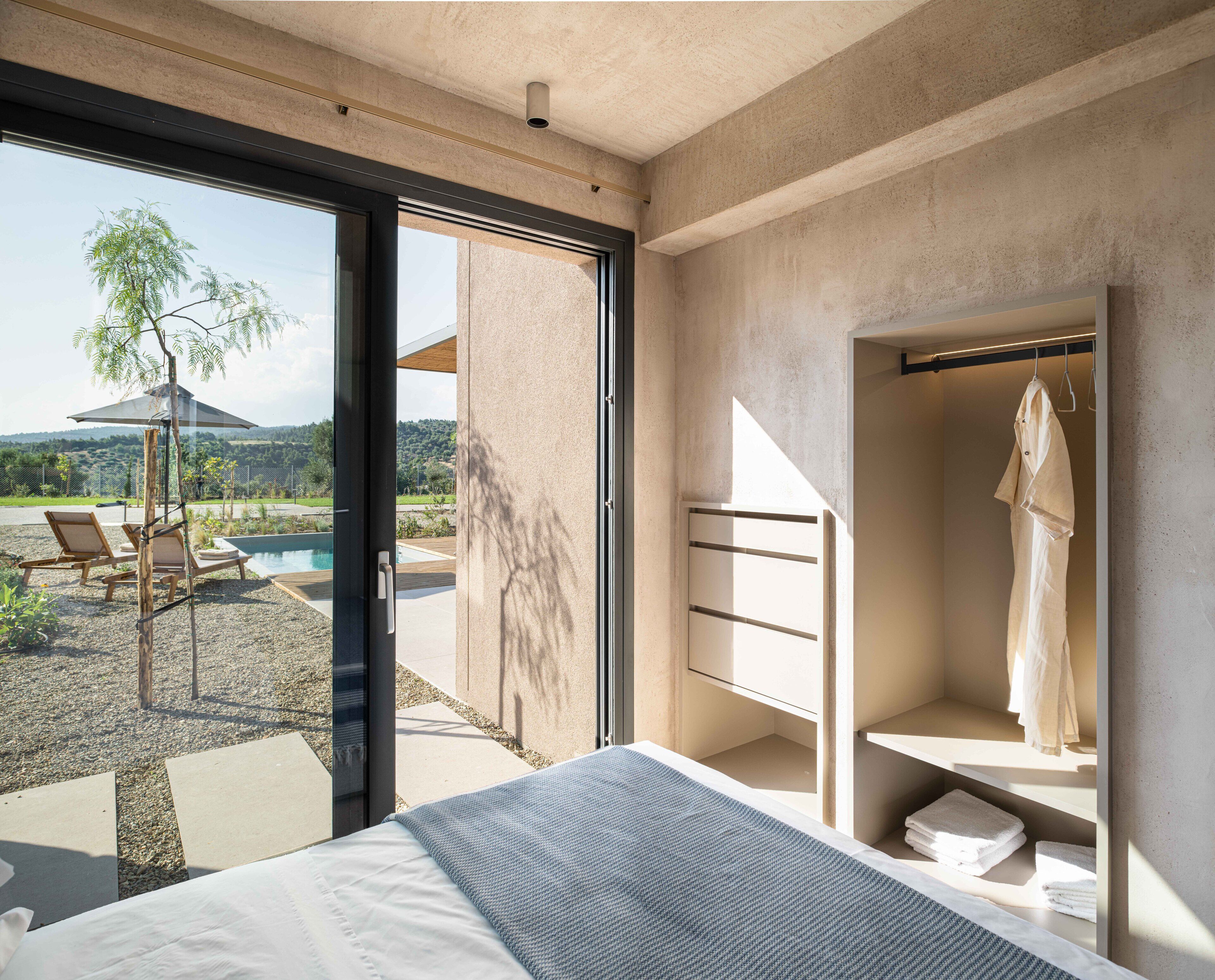 A modern bedroom with a large window, a bed, a walk-in closet, and a view of a pool and garden.
