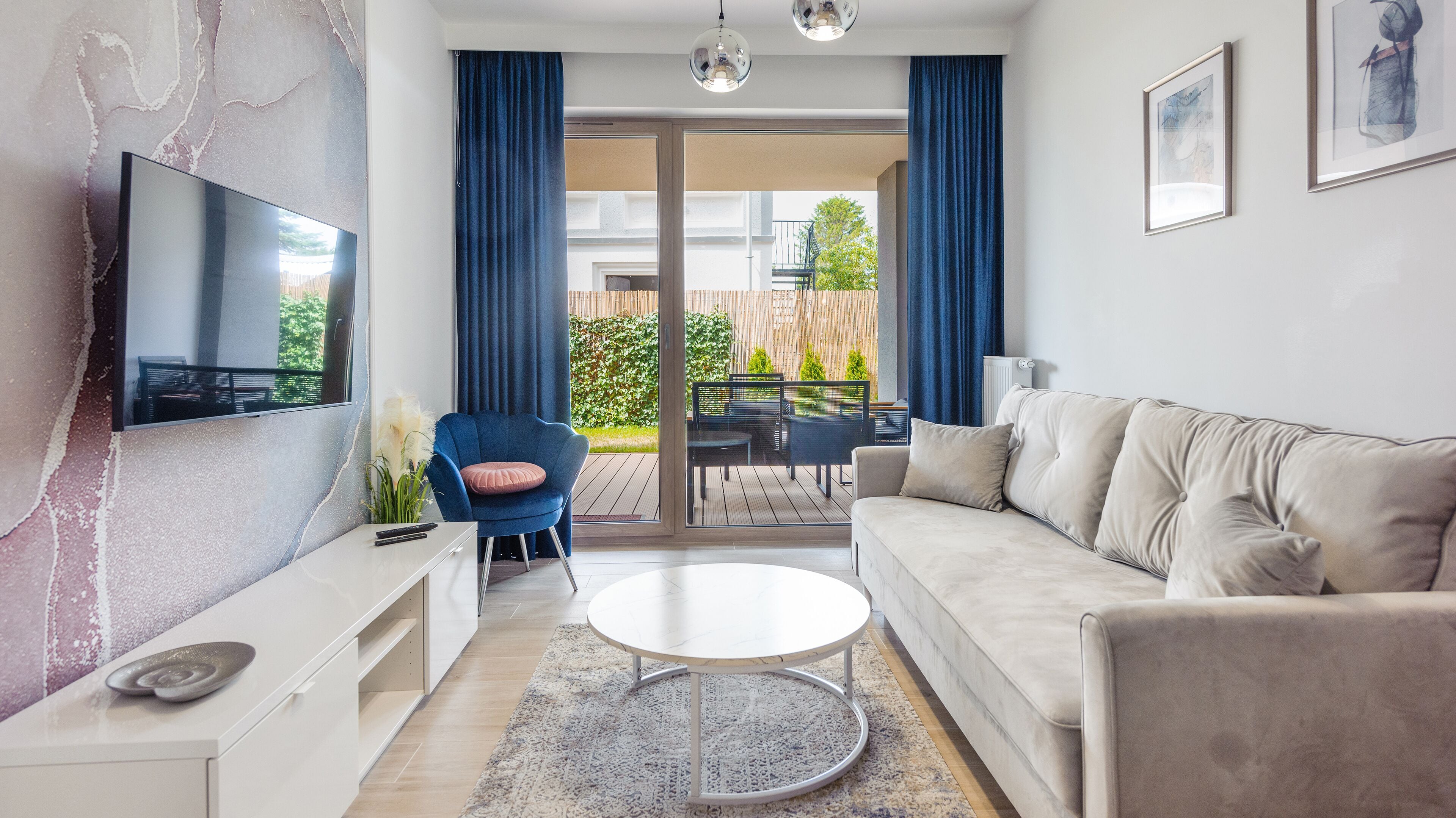 A modern living room with a flat-screen TV, a sofa, a coffee table, and a chair. There is a balcony with a view of greenery and a building.