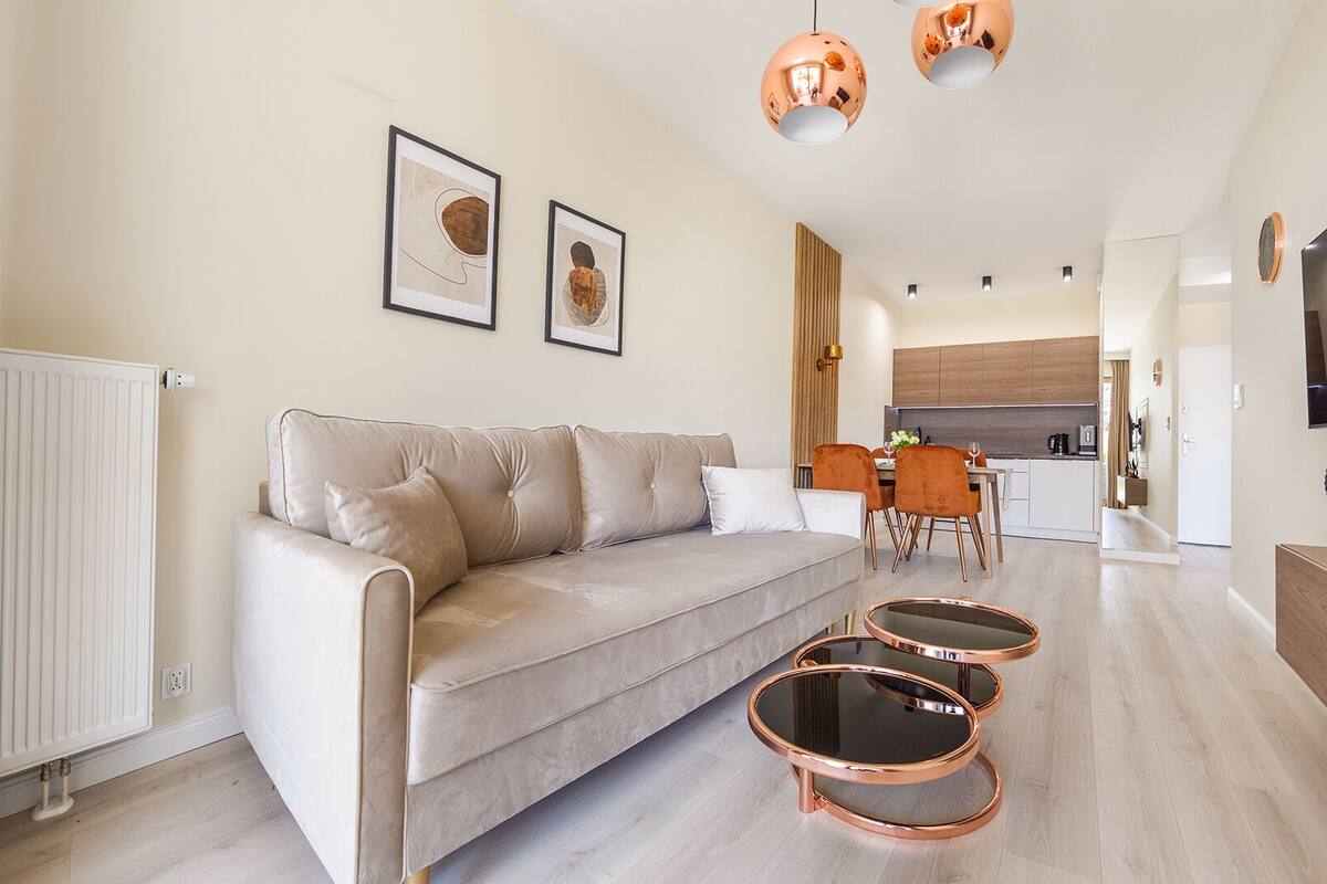 A modern living room with a sofa, two round coffee tables, and a dining area with a table and chairs. There is a kitchen in the background with a TV mounted on the wall.