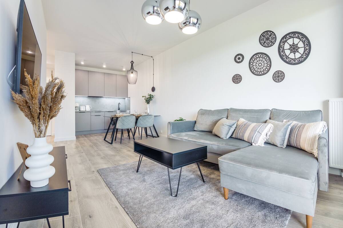 A modern living room with a grey sectional sofa, a black coffee table, and a dining area with a kitchen in the background.