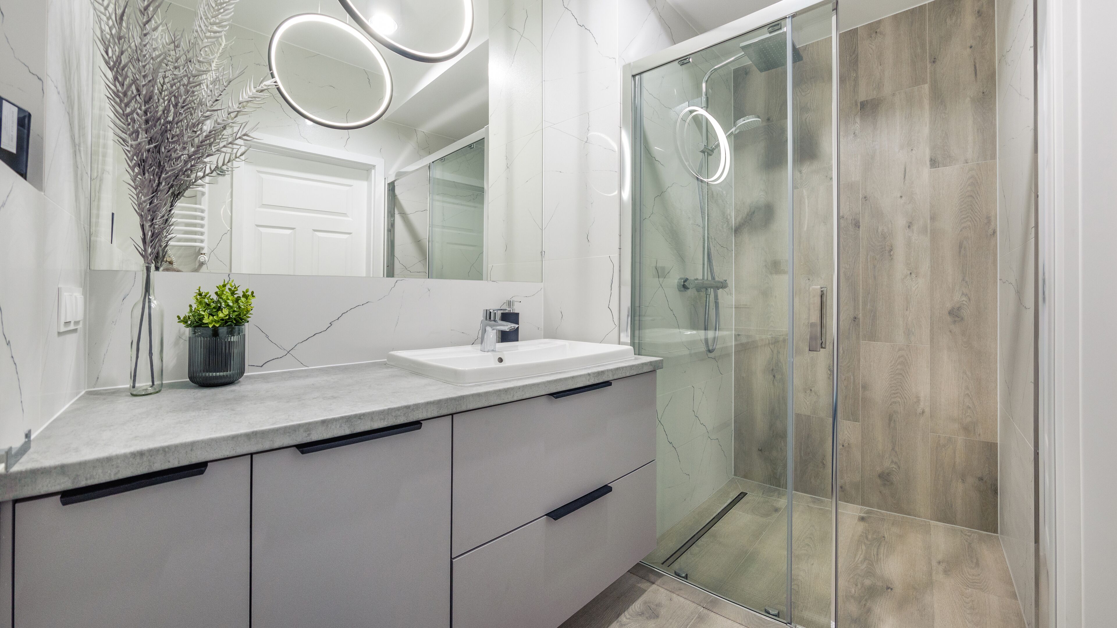 A modern bathroom with a glass-enclosed shower, a white marble vanity, and a large mirror with circular lights.