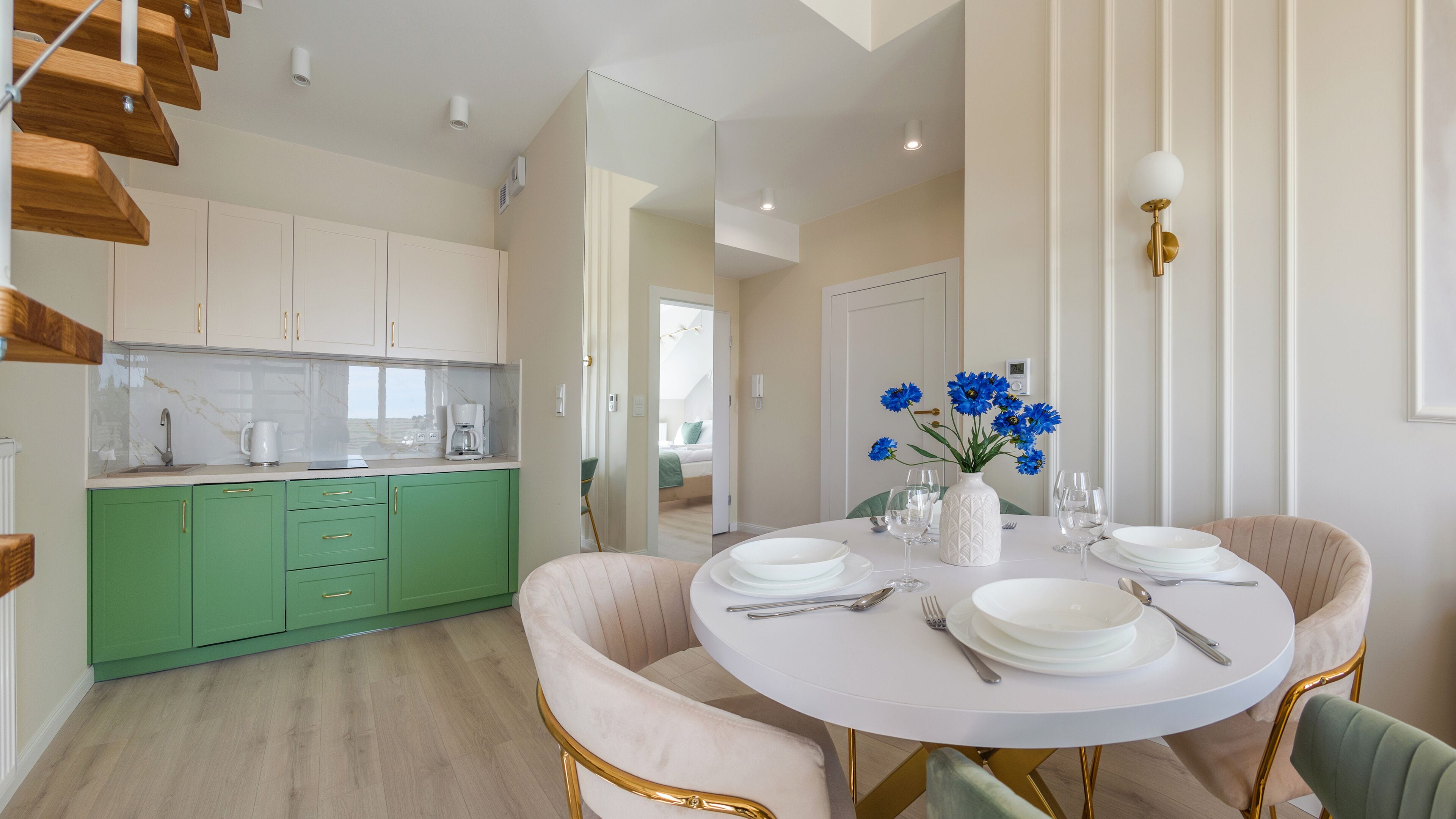 Une cuisine moderne avec des meubles verts, un plan de travail blanc et une machine à café. On y trouve un coin repas avec une table ronde pour quatre personnes, un vase avec des fleurs bleues et un escalier avec des marches en bois.