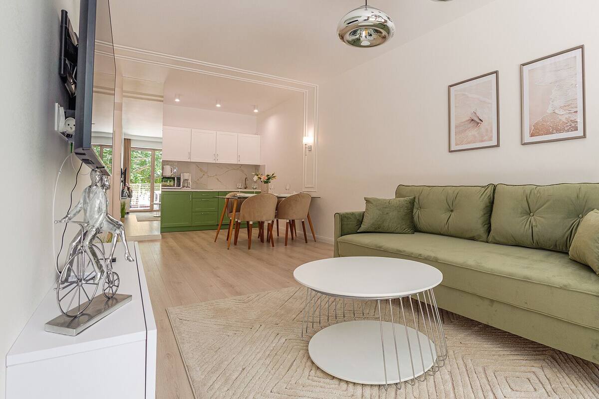 A modern living room with a green sofa, a white coffee table, and a dining area with a green kitchen in the background.