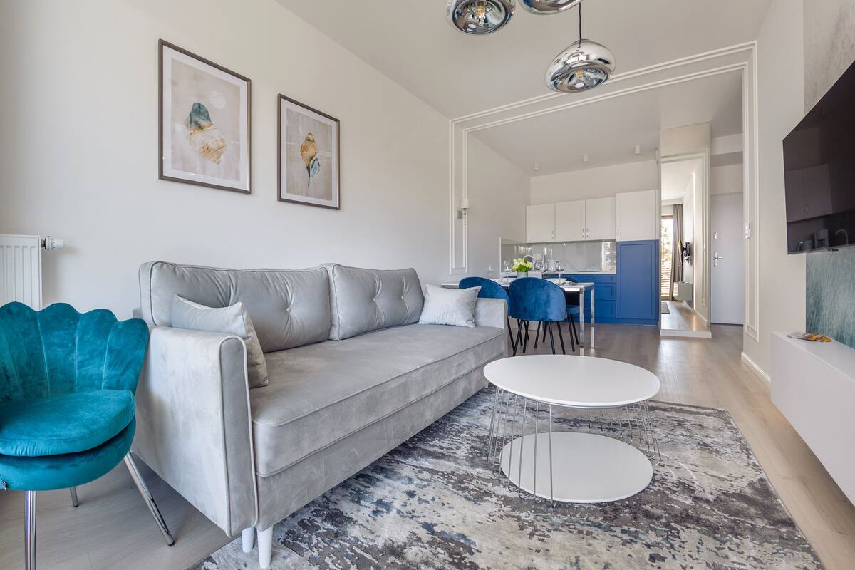 A modern living room with a grey sofa, teal armchair, and a round coffee table. There are two framed artworks on the wall, a kitchen in the background, and a dining area with blue chairs.