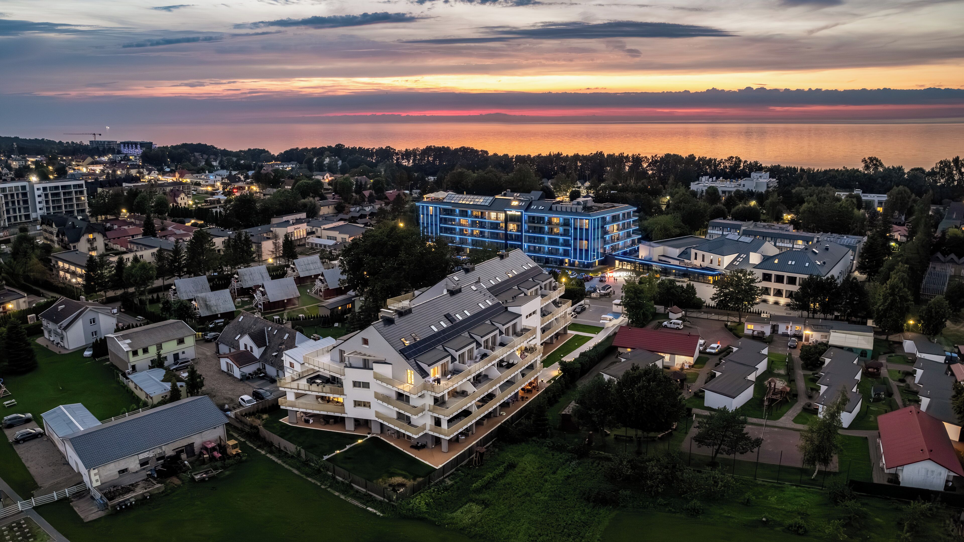 Vue aérienne d’un quartier résidentiel avec des bâtiments modernes, dont une structure imposante illuminée de lumière bleue, entourée de verdure et bordée par un plan d’eau.