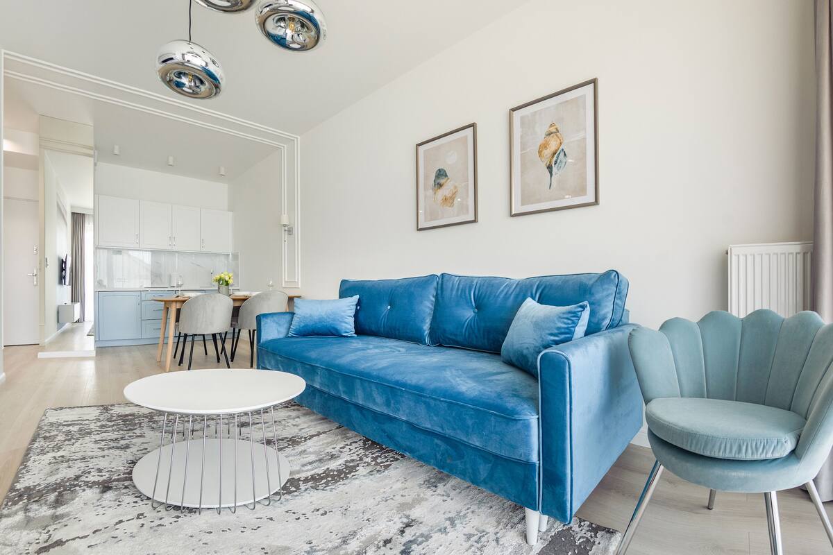 A modern living room with a blue sofa, a round coffee table, and a teal armchair. There is a kitchen in the background with white cabinets and a dining area with a table and chairs.