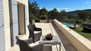 A balcony with wicker furniture, a table with a flower pot, and a view of a swimming pool and greenery. - Beautiful Provencal house with magnificent view of pool and garden (ROGNES)