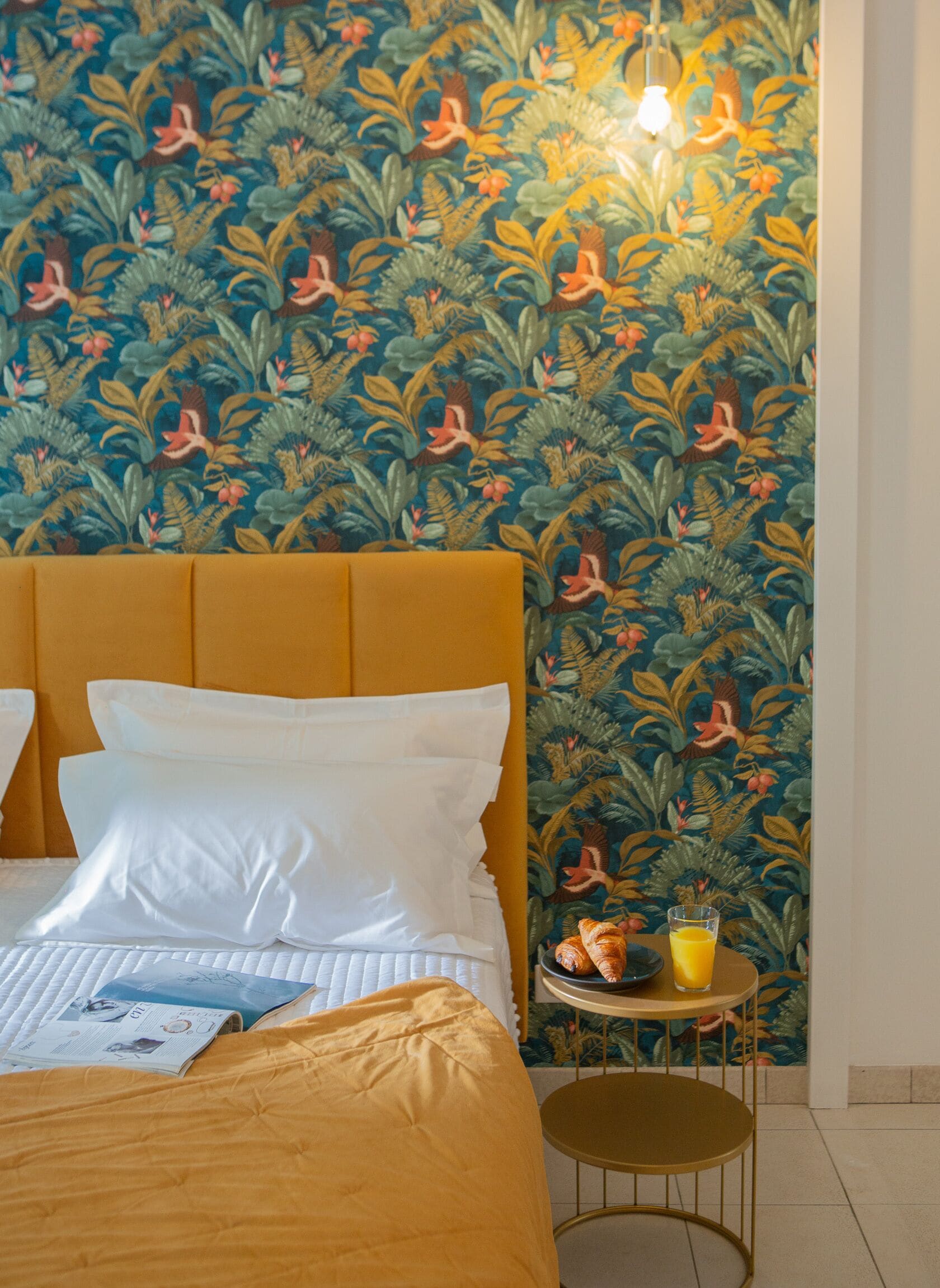 Habitación de hotel con cama, una mesita redonda con una bebida y pared con papel tapiz tropical.