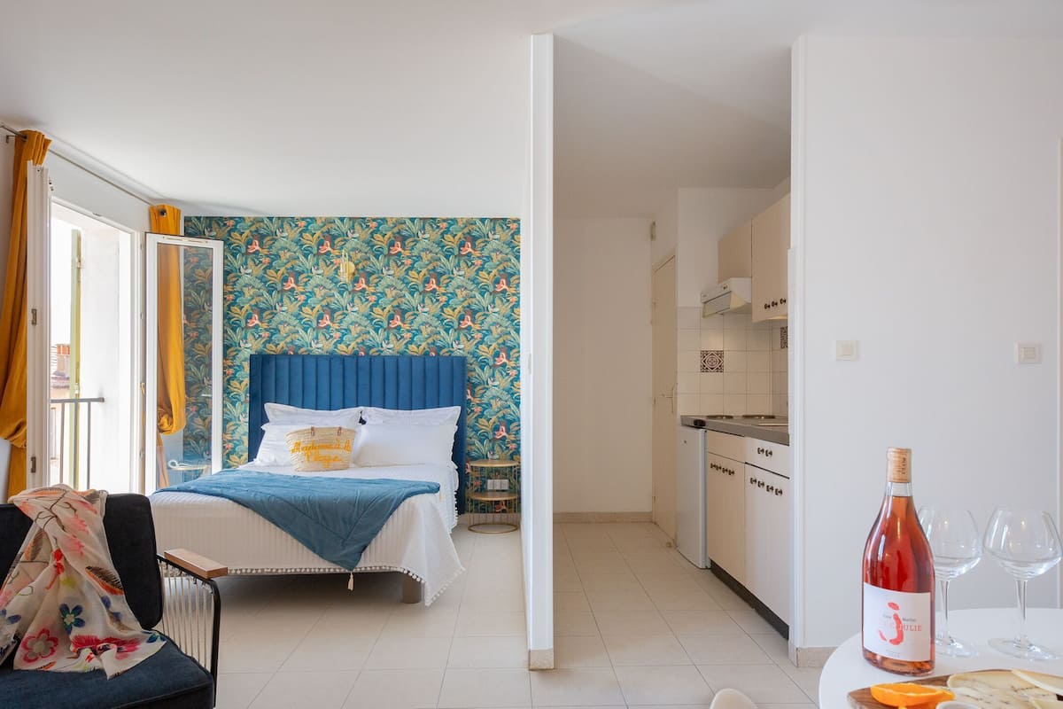 A hotel room with a bed, a small table with a wine bottle and glasses, a kitchen area, and a bathroom with a shower curtain.