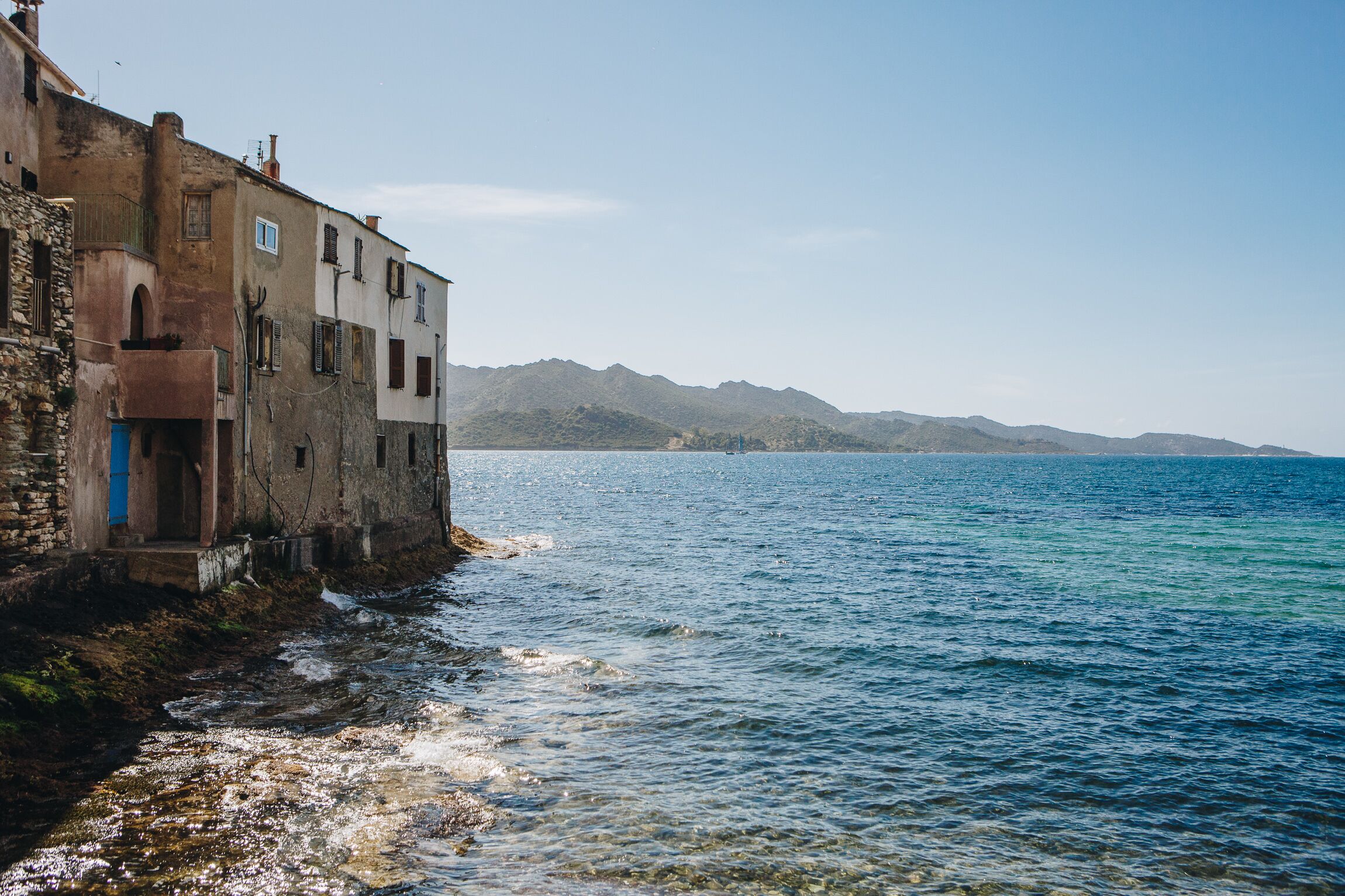 A coastal building with a stone facade, a visible entrance, and a clear view of the sea and distant mountains.