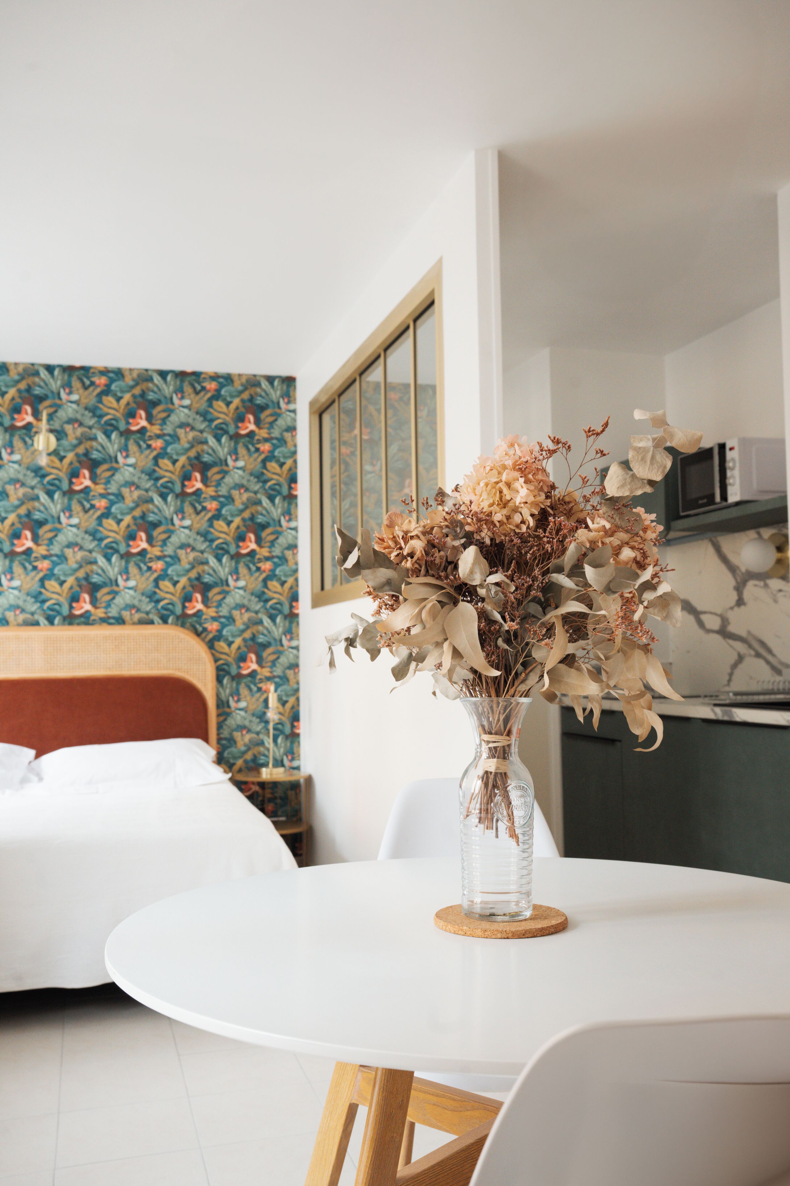 A room with a bed, a dining table with a vase of dried flowers, a patterned wallpaper, and a microwave.