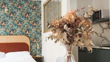 A room with a bed, a dining table with a vase of dried flowers, a patterned wallpaper, and a microwave.